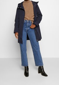 Marinblå parka med huva, beige tröja, högmidjade blå jeans och svarta klackstövlar. Funktioner inkluderar dragkedjestängning och fickor.