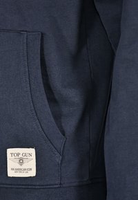 Marineblauwe hoodie met een kangoeroezak. Heeft een gestructureerde stof en een label met "TOP GUN" en "EEN AMERIKAANS ICOON" in het wit.