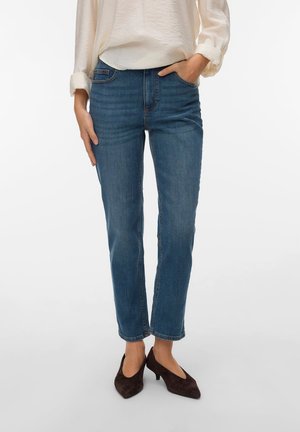 Femme portant un jean droit bleu, un chemisier crème à manches longues et des chaussures marron foncé à talons bas, debout avec une main dans la poche.