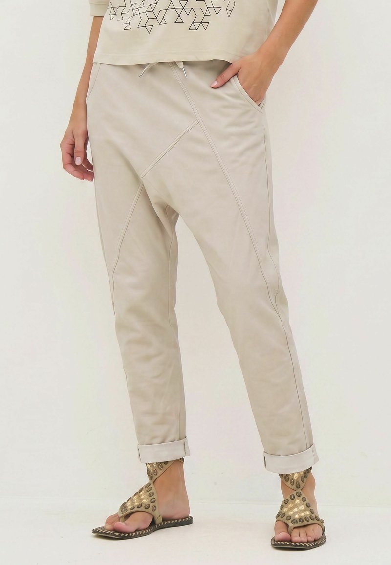 Pantalon beige avec coutures diagonales et revers roulés, porté avec des sandales ouvertes à bouts cloutés dorés et une main dans la poche.