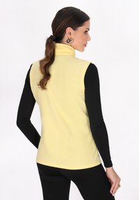 Vest din fleece galben cu un guler înalt, având o textură netedă și un design ajustat, purtat cu un top negru cu mânecă lungă dedesubt.