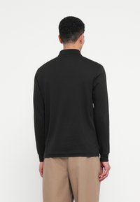 Muž s krátkymi kučeravými vlasmi, ktorý má na sebe čiernu dlhú košeľu a béžové nohavice, stojí otočený chrbtom proti jednoduchému bielému pozadiu.