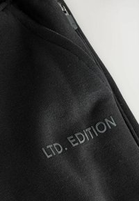 Schwarzer Stoff mit glatter Textur, mit erhabener Aufschrift "LTD. EDITION". Das Design umfasst ein Detail mit einer Reißverschlusstasche.