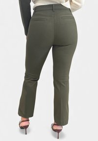 Olivengroene, op maat gemaakte broek met een slanke pasvorm, voorzien van twee achterzakken en een lichte uitlopende zoom, gecombineerd met sandalen met hoge hakken en bandjes.