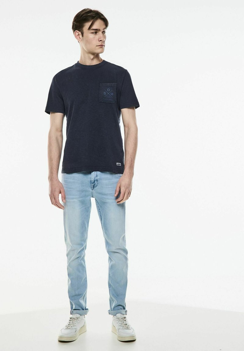 Námořnická modrá bavlněná tričko s kapsou na hrudi a světle modrými akcenty, v kombinaci se světle modrými džíny a bílými teniskami, stojící na neutrálním pozadí.