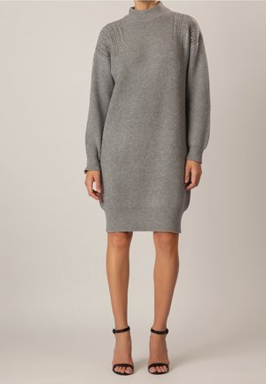Mujer con vestido de punto gris de manga larga y cuello alto, combinado con tacones negros de tiras al tobillo y punta abierta.