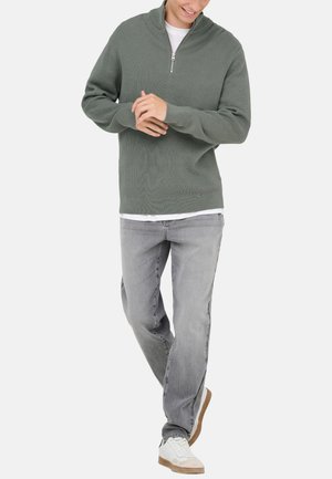 Pullover trøje i dæmpet grøn, med en lynlås i kvart længde og ribbet tekstur. Kombineret med lysegrå jeans og hvide sneakers.