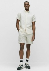 Lichtgekleurde linnen shirt met korte mouwen, gecombineerd met bijpassende shorts. Draagt grijze sneakers en witte sokken met zwarte strepen.