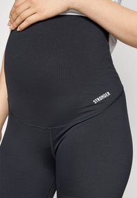 Sorte mammaleggings i svart, laget av elastisk stoff, med høy midje og ordet "STRONGER" trykt på siden i hvitt.