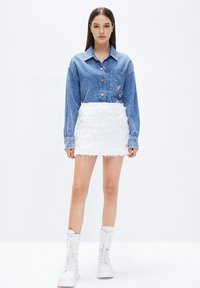 La camicia di denim presenta un colletto classico e una tasca frontale; la mini gonna bianca con frange aggiunge texture; abbinata a stivali alla caviglia bianchi con lacci.
