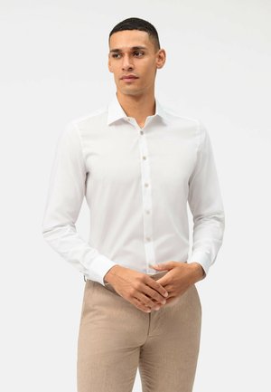 Mężczyzna o krótkich ciemnych włosach, ubrany w dopasowaną białą koszulę zapinaną na guziki oraz beżowe spodnie, stojący na jasnym, jednolitym tle.