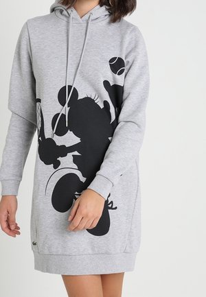 Femme portant une robe à capuche grise avec une grande silhouette noire de Mickey Mouse jouant au tennis, tenant une raquette et une balle.