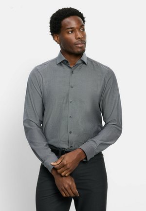 Man with short curly hair wearing a long-sleeve gray button-up shirt and black pants, standing with hands clasped and looking to the side.