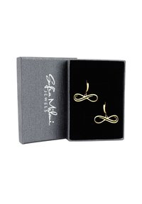 Guld oändlighetssymbol örhängen visade i en grå strukturerad box med svart sammetsinredning. Varumärkeslogotyp synlig på boxens lock.