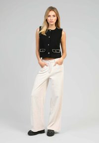 Gilet noir court avec des boutons argentés et des poches, associé à un pantalon large beige clair avec une texture subtile et un léger motif côtelé.