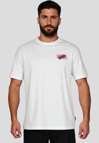 Camiseta blanca de algodón con mangas cortas, escote redondo y texto gráfico rojo en la parte izquierda del pecho. Ajuste estándar y textura suave.