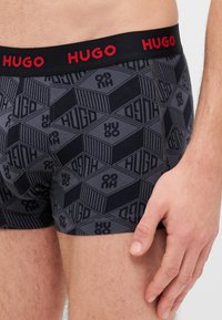 Boxer shorts negros con un patrón de diamantes que presentan el texto "HUGO" en diversas orientaciones. Cintura elástica con letras rojas.