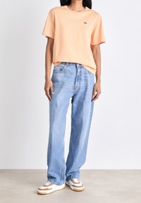 Kvinde iført ferskenfarvet kortærmet t-shirt, lyseblå vide jeans og hvide sneakers med brune detaljer, stående på en ensfarvet gulv.