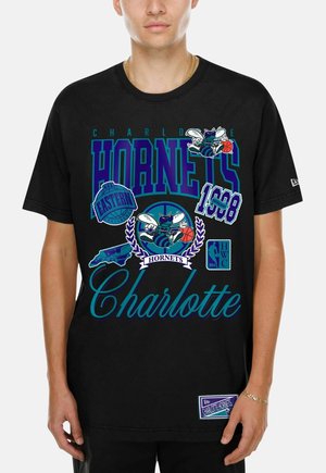 Personne portant un t-shirt noir des Charlotte Hornets avec les logos de l'équipe, du texte et des graphiques en bleu, violet et sarcelle.