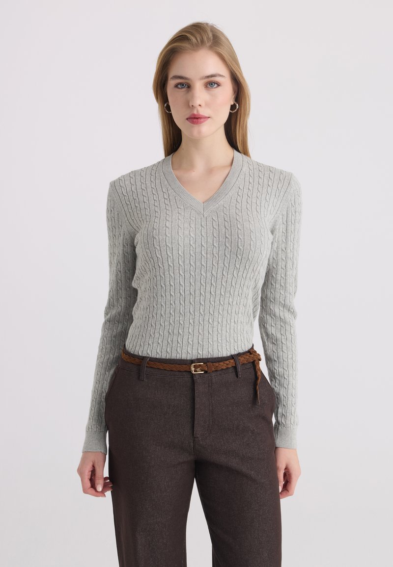 Femme portant un pull gris clair en maille torsadée à col en V, pantalon marron foncé et ceinture tressée marron, debout sur un fond gris clair.