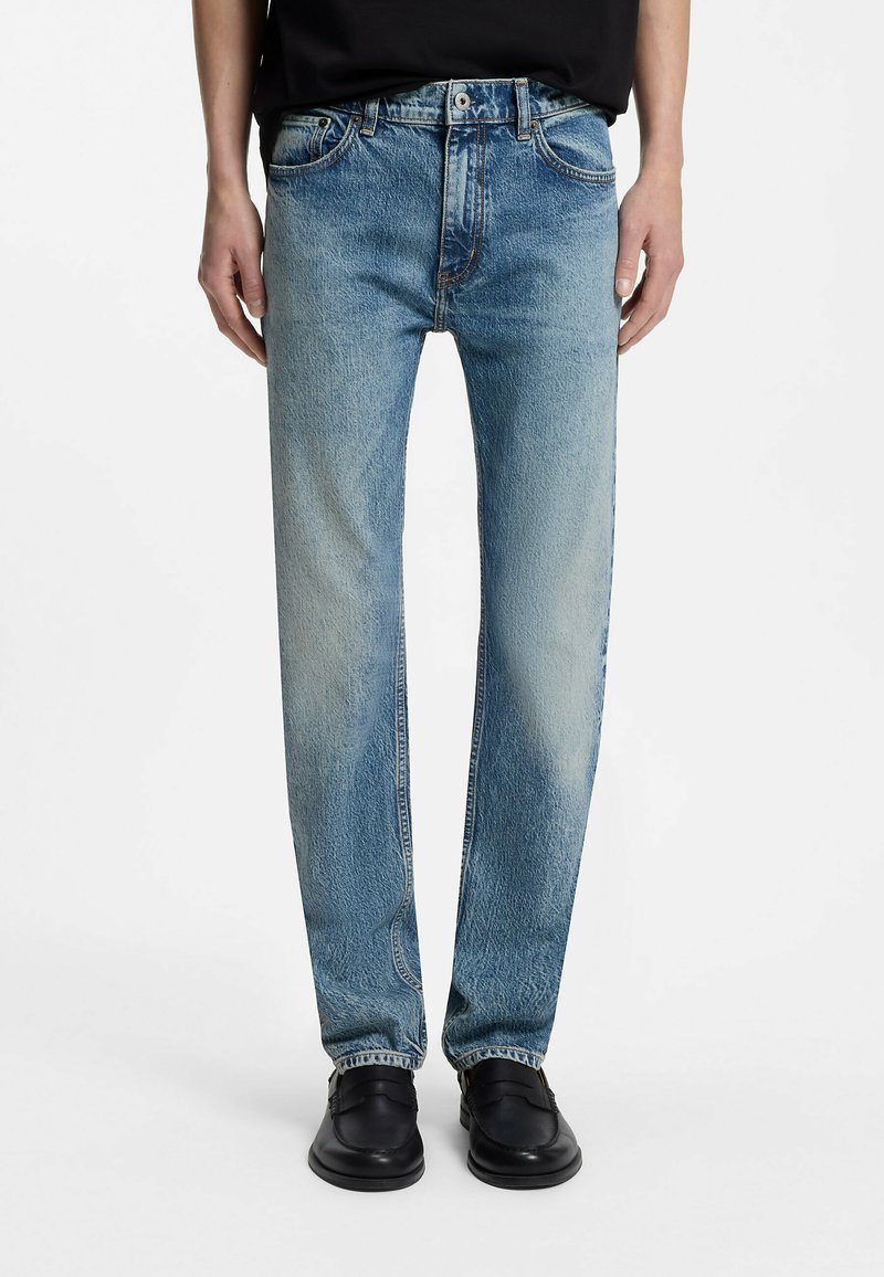 Personne portant un jean droit bleu clair, des mocassins noirs et un haut noir, debout devant un fond blanc.