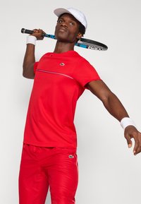 Maglietta sportiva rossa con una striscia orizzontale e logo, abbinata a pantaloni rossi coordinati. Il modello tiene una racchetta da tennis sulla spalla.