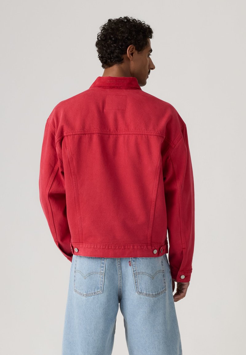Uomo con capelli corti e ricci che indossa una giacca di jeans rossa e jeans azzurri chiari, visto di spalle su uno sfondo semplice.