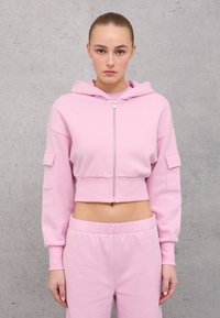 Hoodie cropped rosa chiaro con cappuccio, zip anteriore e tasche a toppa. Texture morbida, polsini a costine e pantaloni della tuta abbinati.
