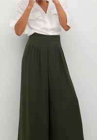 Pantalon large en tissu vert olive avec une taille haute, comportant des plis verticaux et une texture lisse. Associé à une chemise blanche à col.