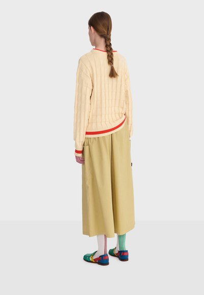 Personne aux cheveux tressés portant un pull crème avec des bordures rouges, un pantalon large beige, des chaussettes rayées dépareillées et des sandales multicolores, vue de dos.