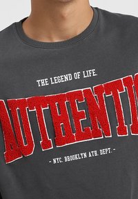 Šedá mikina s kulatým výstřihem, s červeným texturovaným nápisem "AUTHENTIC" a bílým textem nahoře a dole, zdůrazňující atletické téma Brooklynu.