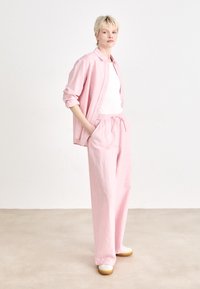 Light pink linen shirt and matching trousers, relaxed fit with a drawstring waist. White tank top underneath, paired with white and tan footwear.