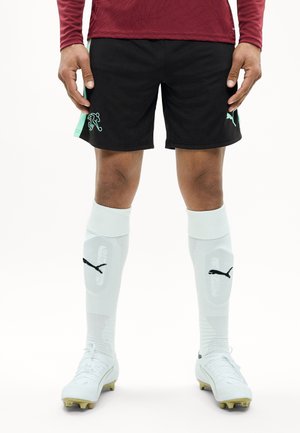 Joueur de football masculin portant un short noir Puma, des chaussettes blanches Puma montant jusqu'aux genoux avec protège-tibias, et des crampons blancs, debout les mains détendues le long du corps.