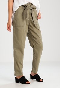 Kvinna i olivgröna högmidjade papperspåsbyxor med självknutbälte, vit topp och svarta öppna klackskor, stående.