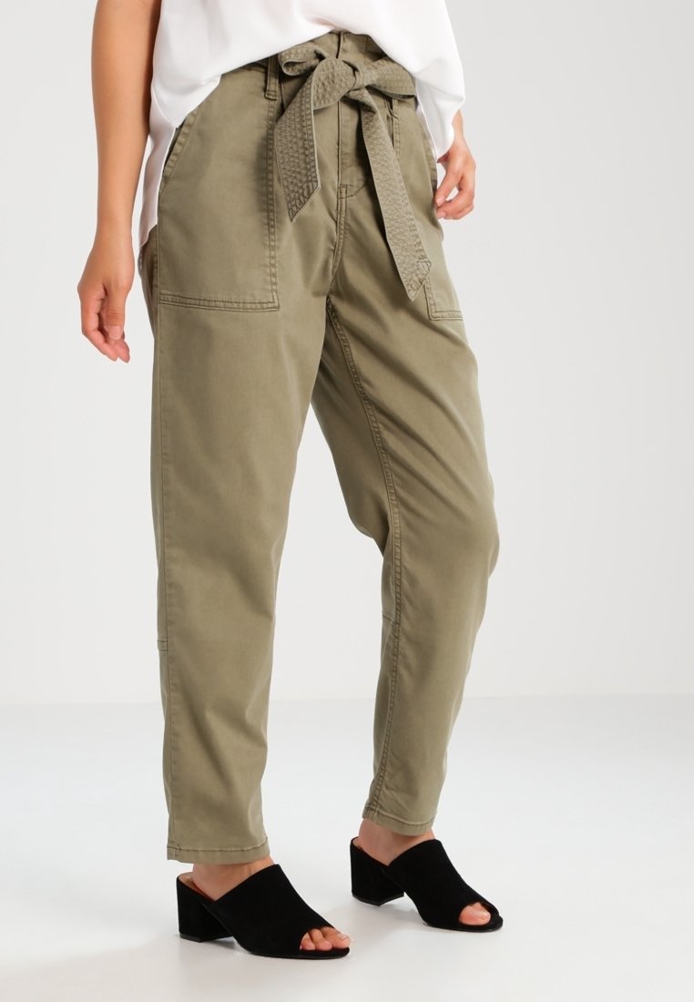 Kvinna i olivgröna högmidjade papperspåsbyxor med självknutbälte, vit topp och svarta öppna klackskor, stående.