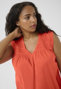 Top sans manches en V, de couleur corail lumineux, avec des détails froncés au niveau du col et une coupe ample et fluide, réalisé dans un tissu au toucher lisse.