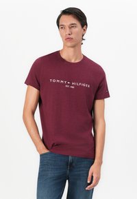 Joven con una camiseta ajustada color burdeos de Tommy Hilfiger con logo y jeans azules, de pie con una mano en el bolsillo contra un fondo blanco.