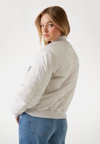 Bomber jacket color beige chiaro con polsini e orlo a coste, caratterizzato da un sottile motivo e tasca con zip sulla manica. Texture liscia e lucida.