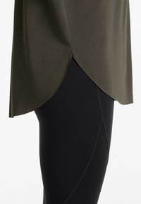 Olivenfarvet top med draperet, asymmetrisk sømad, parret med sorte leggings, der har buede sømdekorationer. Glat tekstur hele vejen igennem.