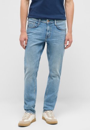 Mann trägt hellblaue Jeans, dunkelblaues Hemd und beige-weiße Sneakers, steht vor einem einfarbigen hellgrauen Hintergrund.