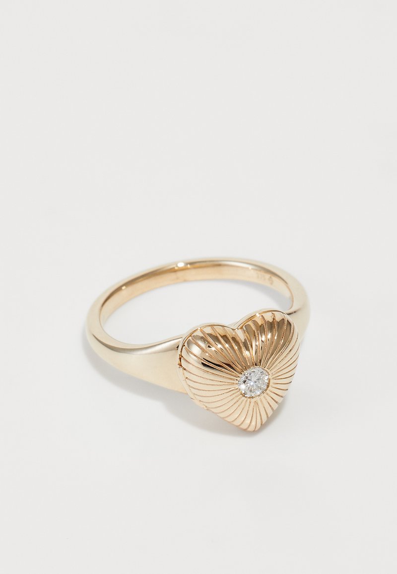 Zlato-obarvan prstan z teksturiranim dizajnom srca, ki ima radiralne linije in osrednji prozoren kamen. Gladek trak z poliranim zaključkom.