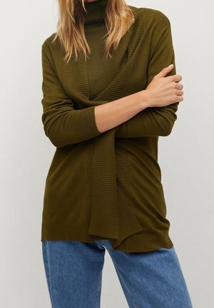 Femme portant un pull en maille côtelée vert olive avec un col roulé et un jean bleu, les bras croisés sur la poitrine.