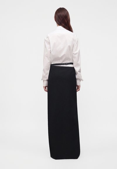 Femme aux longs cheveux bruns vue de dos portant une chemise blanche à manches longues et une jupe longue noire taille haute.