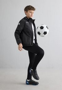 Jaunas berniukas juodos sportinės aprangos viduje laviruoja juodai baltą futbolo kamuolį priešais paprastą šviesią sieną.