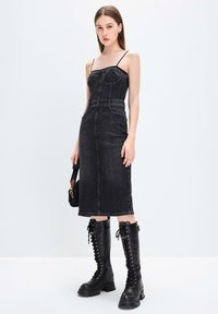 Vestido midi de denim negro con tirantes finos, corpiño estructurado y bolsillos laterales, combinado con botas negras de cordones hasta la rodilla.