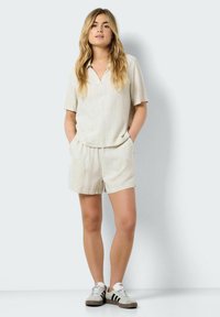 Gai iedegts blakus uzvalks ar īsām piedurknēm un šortiem, izgatavots no strukturētas auduma, ar apkakli un sānu kabatām, kopā ar pelēkiem sporta apaviem.