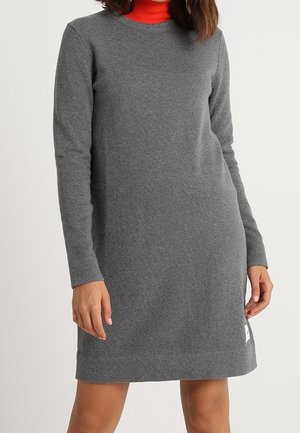 Femme portant une robe en maille grise à manches longues par-dessus un col roulé rouge, debout, les bras détendus le long du corps.