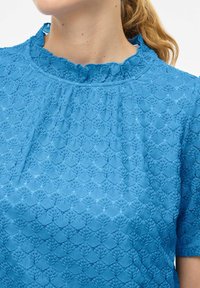 Haut bleu texturé avec un col délicat à festons et une broderie complexe en forme de feuilles. Les manches courtes complètent le design.