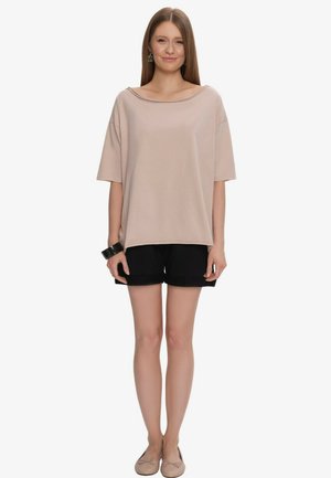 Młoda kobieta stoi, ubrana w luźny beżowy krótki top, czarne szorty, beżowe baleriny, czarną bransoletkę i kolczyki, uśmiechająca się z prostymi włosami.