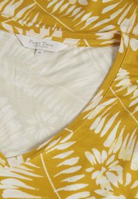 Lyhythihainen paita V-pääntiellä, jossa on sinapinkeltainen tausta, valkoinen lehtikuosi ja pehmeä puuvillan sekoitekangas. Takin merkki on "Part Two."